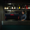 Barista operating a La Cimbali M200 espresso machine in a modern coffee bar with a stylish backlit shelf of liquor bottles