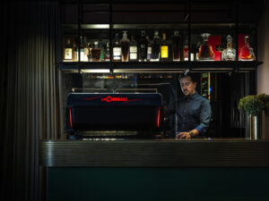 Barista operating a La Cimbali M200 espresso machine in a modern coffee bar with a stylish backlit shelf of liquor bottles